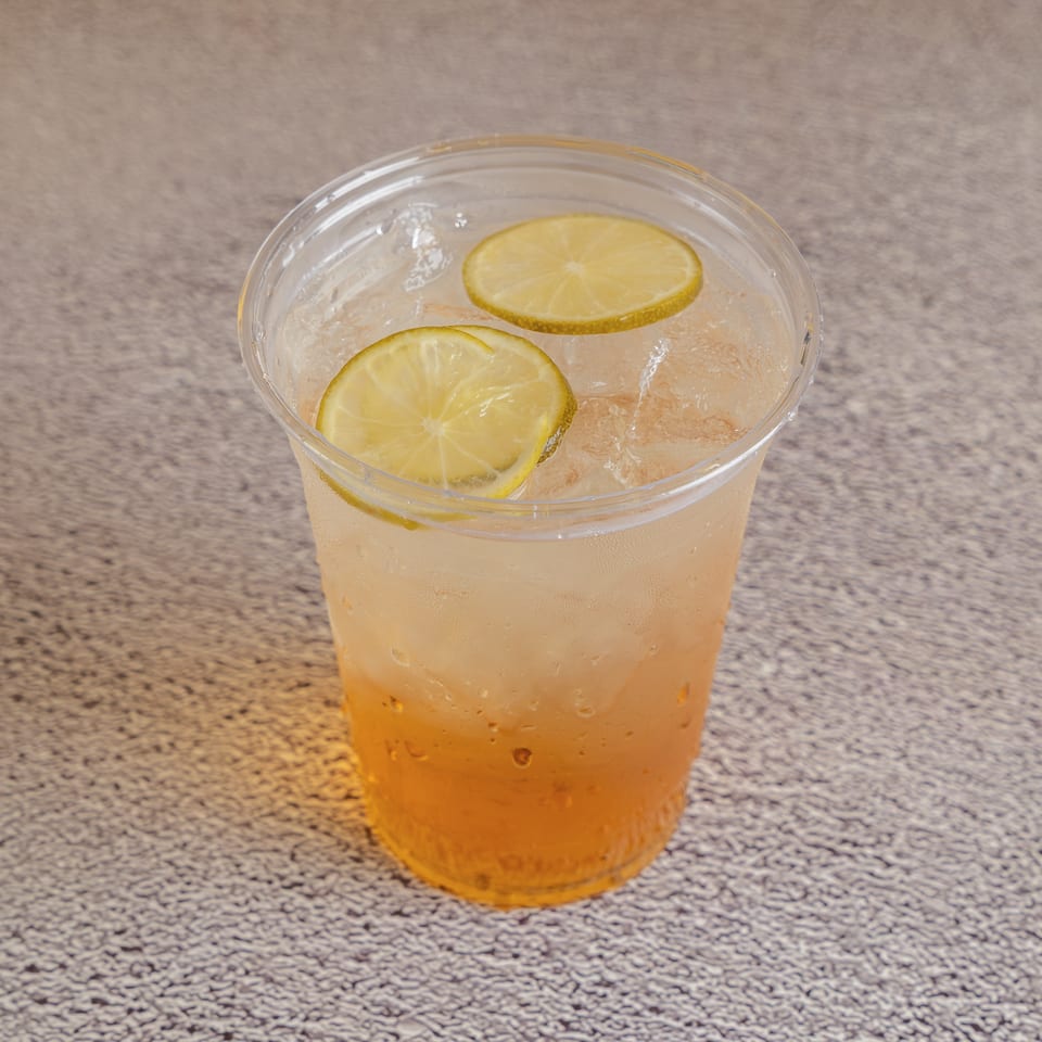Clear plastic cup filled with an iced amber beverage and topped with two lime slices on a speckled countertop.