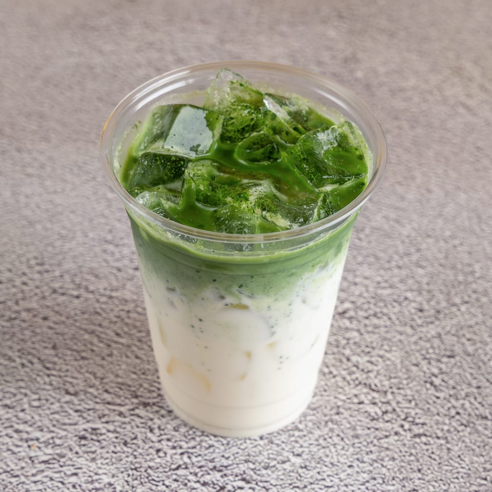 Iced green matcha latte layered over milk in a clear plastic cup on a speckled countertop.